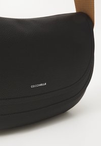Sort læder crossbody taske med en buet form, der har tonale stikninger og et sølvlogo. Stroppen er i en kontrasterende lys brun farve.