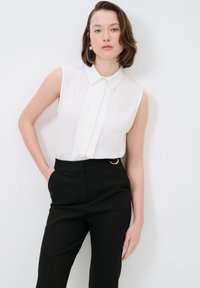Witte mouwloze blouse met een knoopkraag en een plooien aan de voorkant, gecombineerd met zwart hoge taillebroek met een gouden ringaccent.