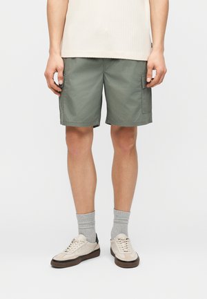 Άνδρας που φοράει χακί cargo σορτς, γκρι κάλτσες μέχρι τον αστράγαλο, και μπεζ αθλητικά παπούτσια, στέκεται μπροστά σε απλό ανοιχτό φόντο.