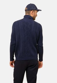 Navyblauer Strickpullover mit hohem Kragen und kompliziertem Zickzackmuster. Weiche Textur, figurnaher Schnitt, kombiniert mit passenden dunklen Hosen.