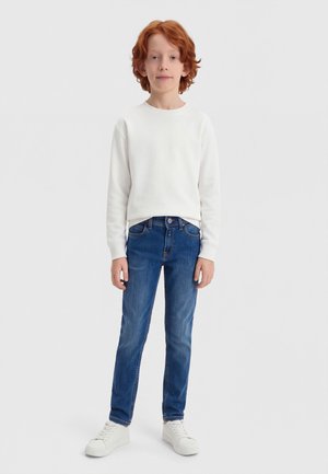 Kind mit roten lockigen Haaren, das ein weißes langärmeliges Shirt, blaue Jeans und weiße Turnschuhe trägt, steht vor einem schlichten hellen Hintergrund.