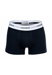 Boxer in cotone blu navy con elastico bianco in vita che mostra la scritta "LACOSTE" in lettere nere. Presenta una parte anteriore sagomata e dettagli di cucitura.