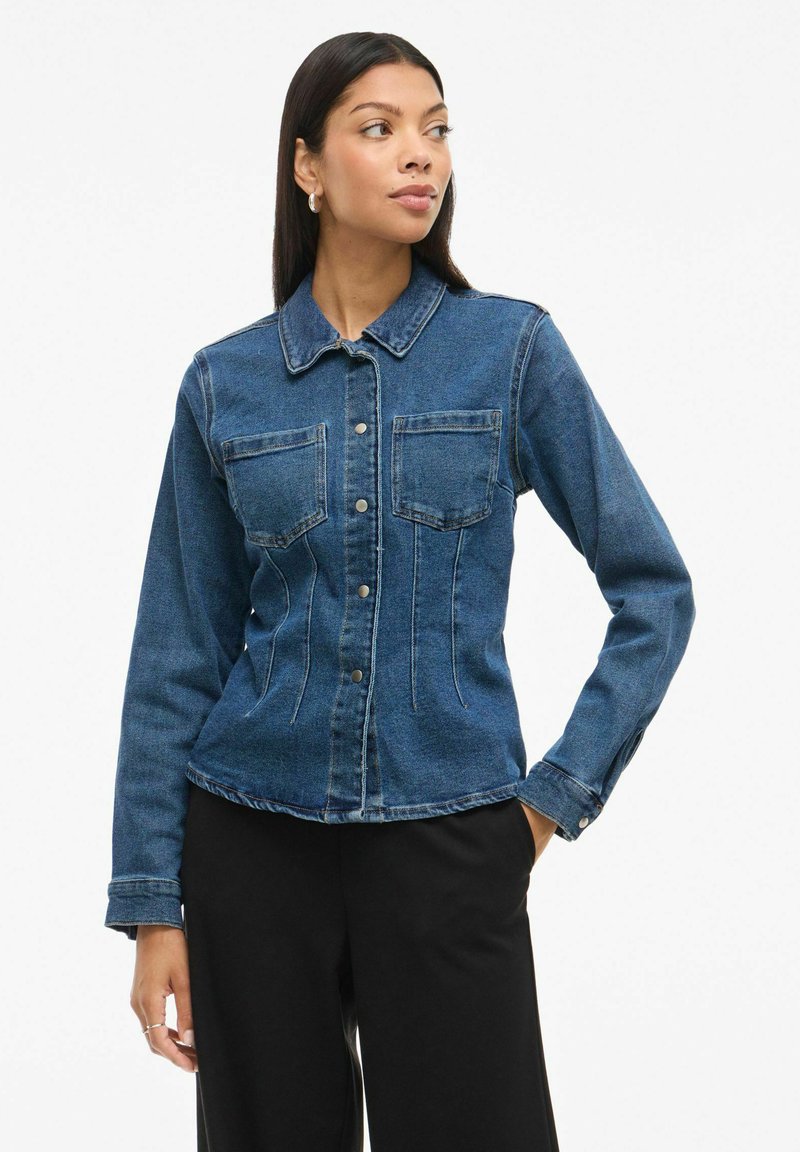 Jeans jakna s kragnim izrezom, sprednjimi žepi, zapiranjem na gumbe in prilegajočim krojem. Ima temno modro barvo z blagim zbledelostjo.