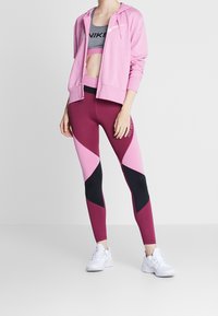 Sudadera rosa con cremallera sobre un sujetador deportivo gris. Los leggings tienen un diseño en bloques de color en burdeos, rosa y negro. Zapatillas deportivas blancas.
