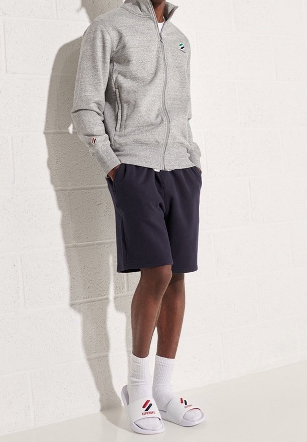 Personne portant une veste grise zippée, un short foncé, des chaussettes blanches et des sandales blanches, debout contre un mur clair texturé, les mains dans les poches.