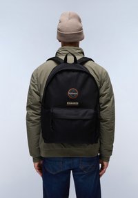 Svart tygryggsäck med rundad form, som har en framficka och logopatch, bärs av en person i en olivgrön jacka och jeans.