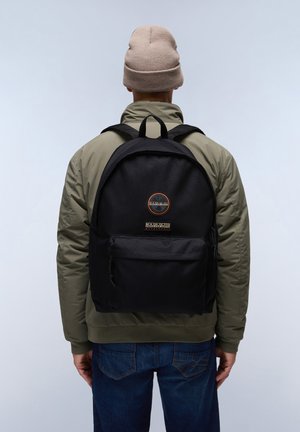 Schwarzer Stoff-Rucksack mit runder Form, ausgestattet mit einer Fronttasche und einem Logo-Patch, getragen von einer Person in einer olivgrünen Jacke und Jeans.