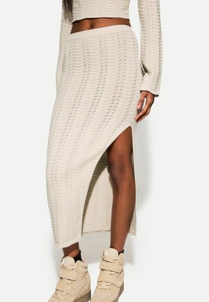 Kobieta ubrana w kremową, dzianinową spódnicę midi z bocznym rozcięciem, w komplecie z dopasowaną bluzką z długim rękawem oraz beżowymi wysokimi trampkami.