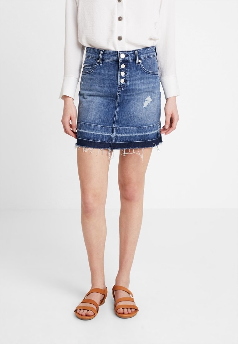 Mini-jupe en denim avec un ourlet effiloché, un lavage bleu clair, une fermeture devant à cinq boutons et des accents usés, portée avec des sandales marron.