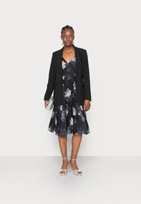 Blazer preto sobre um vestido de comprimento até o joelho com estampado floral em tons escuros, apresentando saia rodada, decote em V e saltos prateados com tiras.