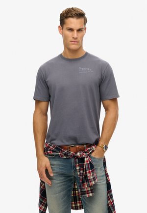 Grijze katoen t-shirt met korte mouwen, met het "Superdry" logo op de borst. Gecombineerd met blauwe jeans en een rood geruit overhemd dat om de taille is geknoopt.