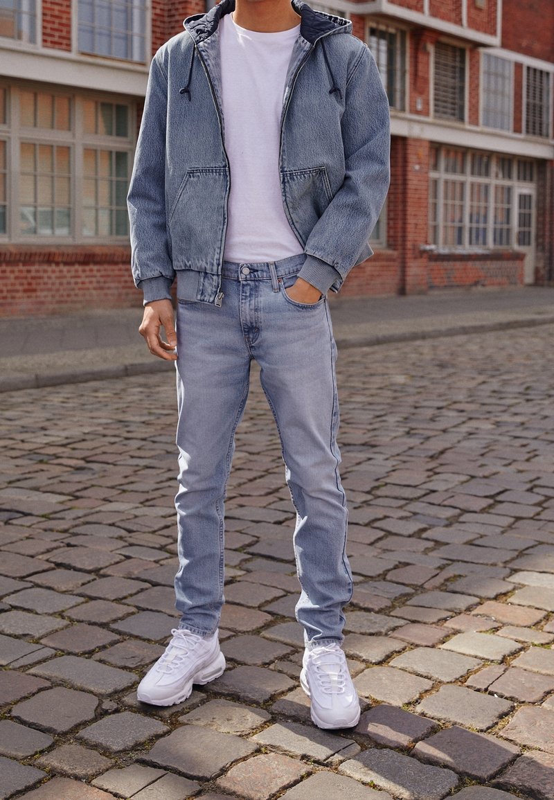 Ljusblå jeansjacka med hood, vit t-shirt och ljusblå jeans i kombination med vita sneakers. Står på kullersten.