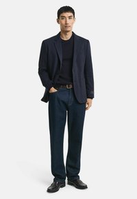 Marinblazer über einem dunklen Pullover, kombiniert mit blauen Jeans und schwarzen Lederschuhen. Der Blazer hat ein einreihiges Design.
