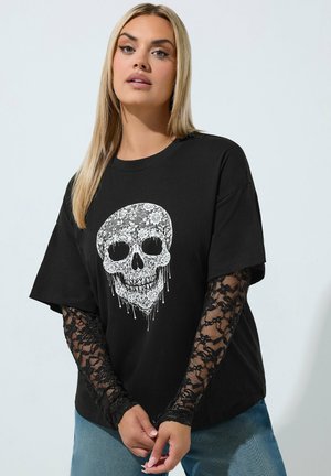 Vrouw met lang blond haar, die een zwarte t-shirt draagt met witte bloemige schedelprint en zwarte kanten lange mouwen, staand tegen een lichte achtergrond.