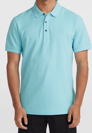 Mann trägt hellblaues Polohemd mit kurzen Ärmeln und schwarzen Knöpfen, steht vor einfarbig hellem Hintergrund.