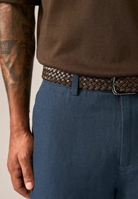Pleteni smeđi kožni pojas nošen preko tamnoplave hlače glatke teksture. Vidljiva srebrna kopča i deo smeđe košulje iznad.