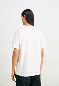 Camiseta de algodón color beige claro, con mangas cortas, cuello redondo y un corte relajado, vista desde la parte posterior.