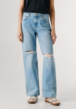 Persona con vaqueros rotos de pierna ancha de color azul claro y zapatos blancos con manchas marrones, de pie contra un fondo liso.