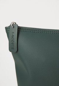 Tamnozeleni teksturirani džep s patentnim zatvaračem. Sadrži oznaku s utisnutim natpisom "LACOSTE". Materijal se čini izdržljivim i laganim.