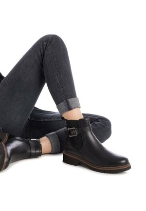 Botas de cuero negras hasta el tobillo con un acento de hebilla, panel lateral texturizado y un tacón bajo, combinadas con jeans oscuros de mezclilla remangados.