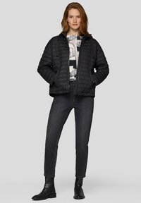 Frau trägt eine schwarze gesteppte Jacke über einem gemusterten Oberteil, dunkle enge Jeans und schwarze Stiefeletten, stehend vor einem hellgrauen Hintergrund.