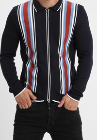 Gilets zippé bleu marine avec des rayures verticales rouges, bleu clair et blanches. Poignets et col côtelés avec une coupe ajustée.