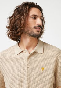 Polo beige avec un design texturé en gaufre. Présente un col de couleur claire et un petit emblème d'arbre jaune sur la poitrine gauche.