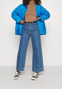 Blaue gesteppte Jacke, brauner Rollkragenpullover, hochtaillierte weit geschnittene blaue Jeans, schwarzer Gürtel und olivgrüne Sneaker mit weißen Akzenten.