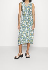 Kukkakuvioinen mekko valkoisena, jossa on vihreitä lehtiä ja keltaisia kukkia, V-pääntiellä ja vyötärölle sidottavalla vyöllä, yhdistettynä mustiin paksupohjaisiin sandaaleihin.