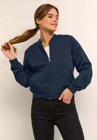 Veste de bombardier marine avec col montant, zip frontal et poignets côtelés, associée à un haut blanc et un jean noir. Texture de tissu lisse.