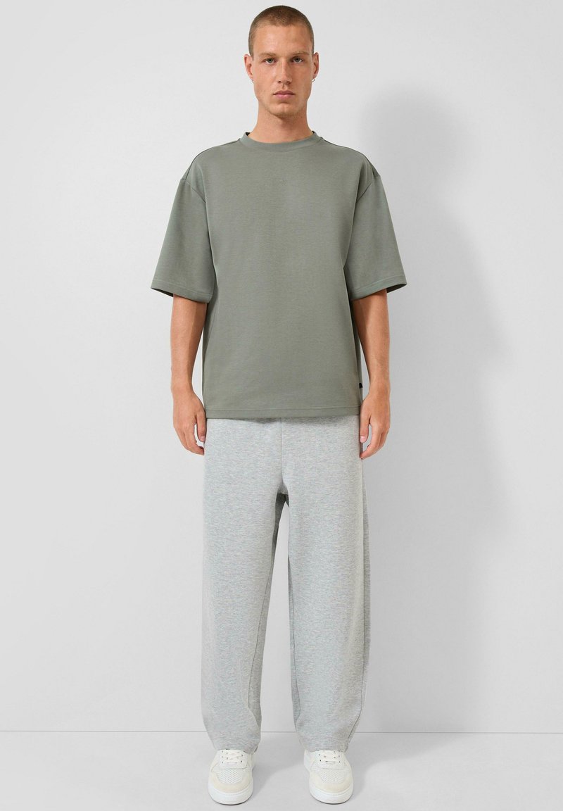 Man in een olijfgroene oversized korte mouwen shirt, lichtgrijze losse joggingbroek en witte sneakers, staande tegen een witte muur.