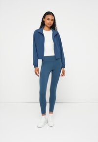 Casaco azul com fecho éclair, camisa branca de gola redonda e leggins teal. Tecido suave, design justo e detalhe de logótipo em contraste nas leggins. Ténis brancos.