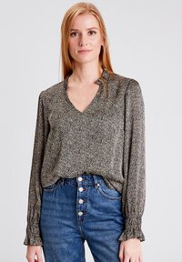 Blusa a motivi in tessuto nero, oro e crema; presenta un collo a V, maniche lunghe con polsi arricciati, indossata con jeans blu a vita alta.