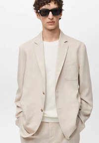 Beige tailored blazer with two buttons and patch pocket, worn over a white knit sweater. Model wearing black sunglasses.