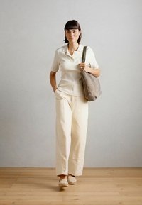 Licht beige geribbelde polo shirt, crèmekleurige brede broek en lichte schoenen; met een zachte grijze tas, tegen een neutrale achtergrond.