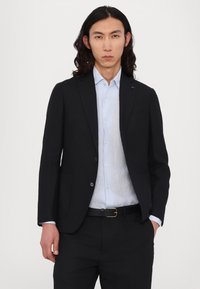Schwarzer maßgeschneiderter Blazer über einem hellblauen Hemd mit Knöpfen, mit sichtbarer Tasche und silbernen Knöpfen. Getragen mit dunklen Hosen und einem schwarzen Gürtel.