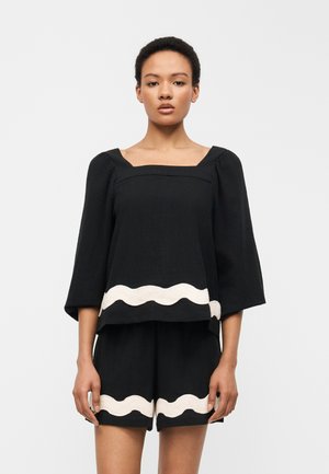 Femme portant un haut noir à encolure carrée et un short assorti avec une bordure ondulée blanche à l'ourlet, debout contre un fond uni.