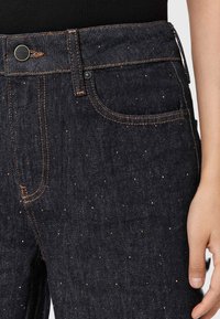 Jeans in denim scuro con pois dorati, design a cinque tasche, cuciture in rame e chiusura con bottone metallico. Texture liscia e forma su misura.