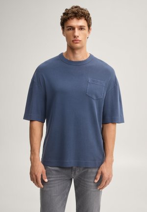 Junger Mann mit lockigem Haar, der ein lockeres blaues T-Shirt mit Brusttasche und graue Jeans trägt, steht vor einem einfarbigen hellen Hintergrund.