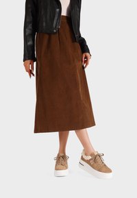 Gonna midi in corduroy marrone abbinata a sneaker in finta pelliccia beige. La giacca è in pelle nera con un design aderente.