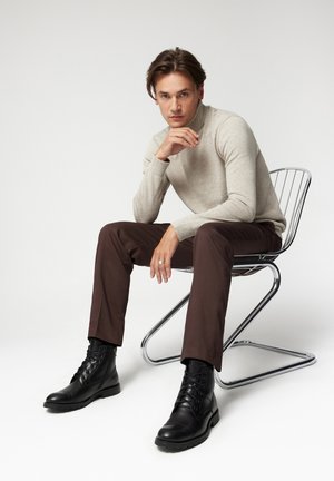 Szary sweter z długim rękawem, brązowe spodnie i czarne buty na sznurówki. Model siedzi na metalowym krześle na neutralnym tle.