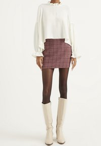 Donna che indossa una blusa bianca con volant, una minigonna a quadretti rossi, collant neri e stivali crema alti fino al ginocchio, in piedi di fronte a uno sfondo semplice.