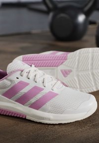 Άσπρα και ροζ παπούτσια τρεξίματος Adidas σε ξύλινο πάτωμα με θαμπά μαύρα kettlebells στο φόντο.