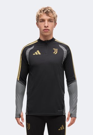 Jeune personne portant un haut de sport à manches longues noir et gris ainsi qu'un pantalon avec les logos dorés de la Juventus et d'Adidas à l'avant.
