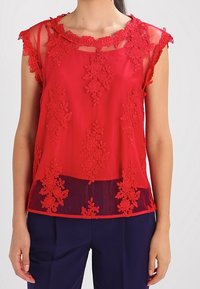 Rød gennemsigtig bluse med detaljeret blomsterlacemønster langs halsudskæringen og kanten, med korte, gennemsigtige ærmer. Parret med mørke bukser.