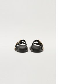Black sandals featuring a flat sole, dual straps, and gold-tone hardware accents. The material appears smooth with a sleek design.