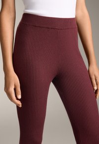 Leggings bordeaux a coste con una vita alta, che mostrano un tessuto a maglia strutturata. La vista ravvicinata rivela cuciture lisce e una forma aderente.