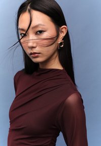 Viininpunainen yläosa, jossa on laskostettu muotoilu ja läpikuultavat hihat, yhdistettynä suureen kultakorvakoruun. Taustana on vaaleansininen. Hiukset laskeutuvat kasvojen yli.