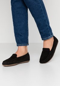 Black suede loafers with a smooth texture, featuring a stitched design and a low rubber sole, worn with dark blue denim jeans.
