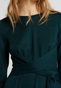 Tmavě modrá obepínací šaty s kulatým výstřihem, dlouhými rukávy a akcentem na vázání v oblasti pasu. Tkanina má jemný, texturovaný vzhled.